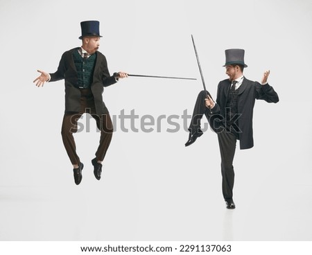 Remembered childhood. Portrait of two men, friends wearing classic suits jumping and fighting with canes over white studio background. Concept of duel, battle, historical remake, comparison eras, ad Royalty-Free Stock Photo #2291137063