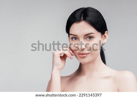 Close-up beauty portrait of young woman with smooth healthy skin, she gently touches her face with her fingers on light grey background and smiles. Skincare concept