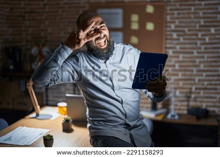 Young hispanic man with beard and tattoos working at the office at night doing peace symbol with fingers over face, smiling cheerful showing victory 