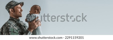 cheerful army soldier in uniform holding newborn son isolated on grey, banner