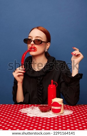 Young beautiful girl eating spicy red chilli pepper and mayonnaise over blue background. Food pop art photography. Complementary colors. Concept of art, beauty, food. Copy space for ad, text.