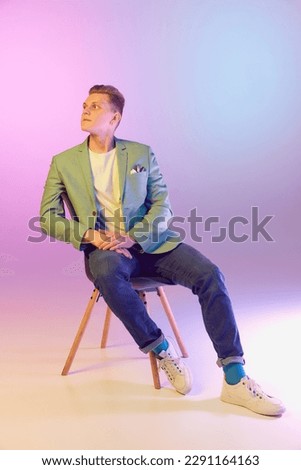 Elegant, casual look. Young handsome man in stylish jacket posing, sitting on chair over gradient purple background in neon light. Concept of emotions, lifestyle, youth, modern fashion. Ad