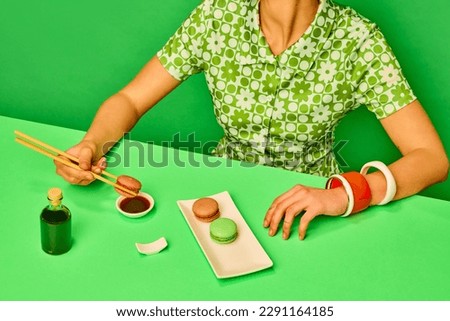 Cropped image of girl putting sweet macarons into soy sauce with chopsticks against green background. Extraordinary taste combination. Food pop art photography. Concept of retro style, creative vision Royalty-Free Stock Photo #2291164185