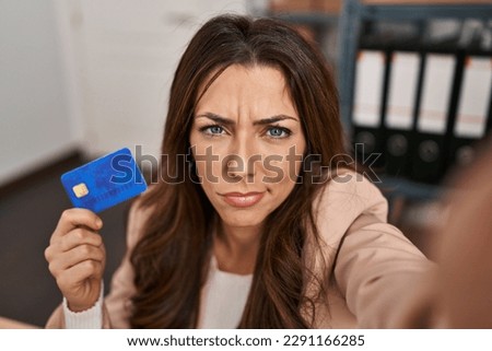 Young brunette woman working at small business ecommerce holding credit card skeptic and nervous, frowning upset because of problem. negative person. 