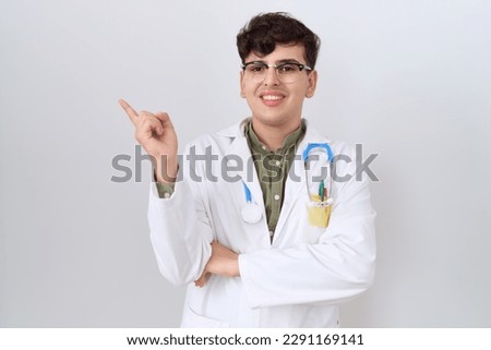 Young non binary man wearing doctor uniform and stethoscope with a big smile on face, pointing with hand and finger to the side looking at the camera. 