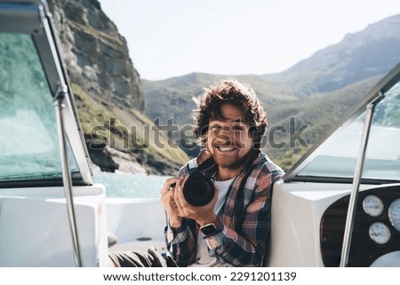 Delighted bearded male photographer in casual wear with professional camera enjoying boat trip through mountains and river on sunny weather