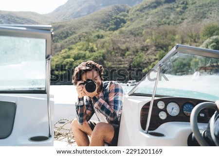 Young male photographer in casual clothes sitting on boat and taking picture with professional digital camera while riding at yacht during trip