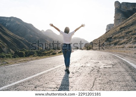 Back view full body of anonymous female traveler in casual clothes raising arms and admiring majestic mountains while standing on empty road Royalty-Free Stock Photo #2291201181