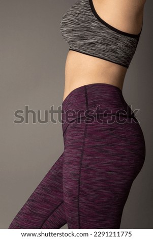 Studio close up photo of a woman wearing purple yoga pants and a sports bra. The background is grey.  Royalty-Free Stock Photo #2291211775
