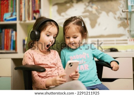 Two little girls sisters wear headphones watching cartoons or kid video on mobile phone. 