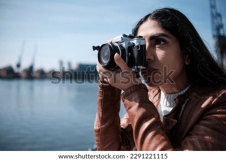 A young woman is taking pictures with film analog camera. There is a lake behind her and she is wearing a leather jacket. No AI concept.