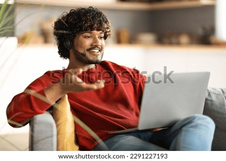 Online Communication. Smiling Indian Man In Headset Making Video Call On Laptop While Sitting On Couch At Home, Happy Young Eastern Guy Talking And Gesturing At Web Camera, Copy Space Royalty-Free Stock Photo #2291227193