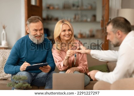 Real Estate Agent Giving New House Keys To Happy Mature Couple Buying Property And Signing Papers Sitting On Sofa Indoors. Apartment Rent And Purchase. Selective Focus On Spouses