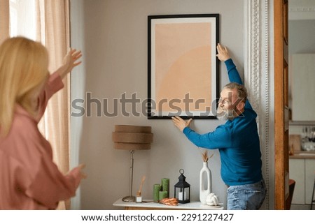 Cheerful Senior Spouses Hanging Poster In Frame On Wall Decorating Modern Living Room Together Standing At Home After Renovation. Interior Design Concept. Selective Focus On Husband