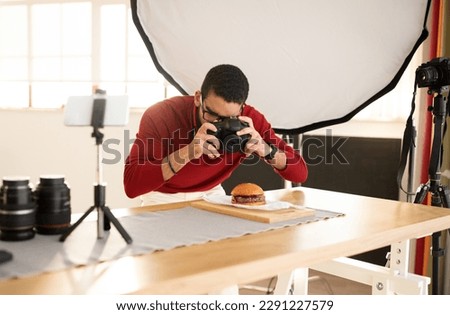 Famous photographer streaming while working on food shoot, young man using digital camera, taking photo of delicious hamburger and recording video on smartphone set on tripod, photo studio interior