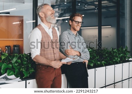 Cheerful male colleagues standing and leaning on potted plants counter in office with glass doors while looking away sharing funny moment and laughing together Royalty-Free Stock Photo #2291243217