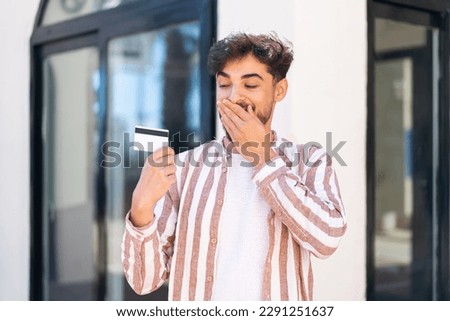 Handsome Arab man at outdoors holding a credit card with surprised expression