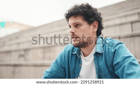 Upset man sits on the stairs and thinks emotionally. Closeup portrait of emotional young bearded man in denim shirt sits on high steps. Royalty-Free Stock Photo #2291258921