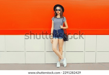 Beautiful woman model posing wearing black round hat, shorts and handbag in the city on orange background