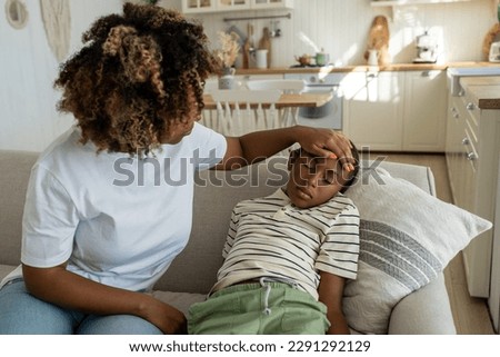 Fever in kids. African American mother touching forehead of sick little boy son at home, caring parent mom checking childs temperature with hand. Schoolboy lying on sofa feeling unwell and ill Royalty-Free Stock Photo #2291292129