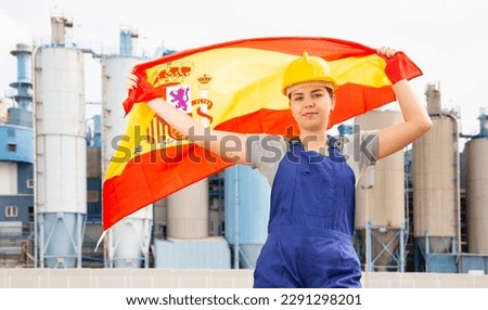 Cheerful female worker in hardhat with spanish flag standing in front of factory