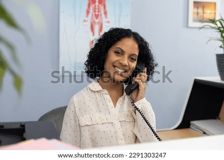 Portrait of happy biracial medical receptionist sitting at reception desk and talking on telephone. Hospital, medicine and healthcare. Royalty-Free Stock Photo #2291303247