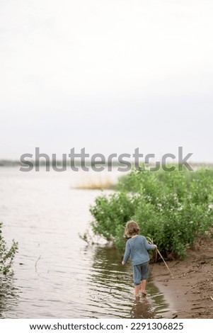 European blond child walks along the sandy beach of the shore of the river and plays with plants