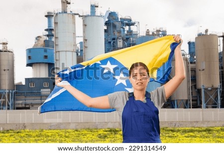 in countryside,worker in overalls,frustrated with working conditions,stands in front of factory building and holds Bosnian flag in her hands