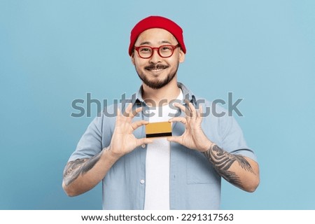Portrait of handsome smiling Asian man wearing stylish red hat and hipster eyeglasses, holding credit card looking at camera isolated on blue background. Shopping, sales concept