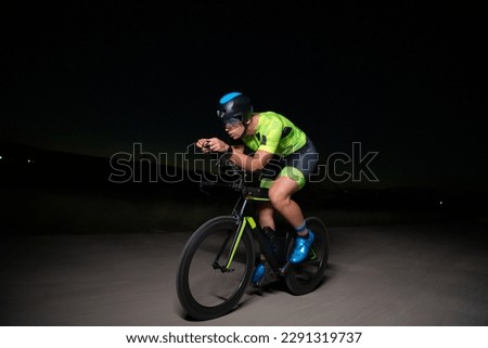 A triathlete rides his bike in the darkness of night, pushing himself to prepare for a marathon. The contrast between the darkness and the light of his bike creates a sense of drama and highlights the Royalty-Free Stock Photo #2291319737