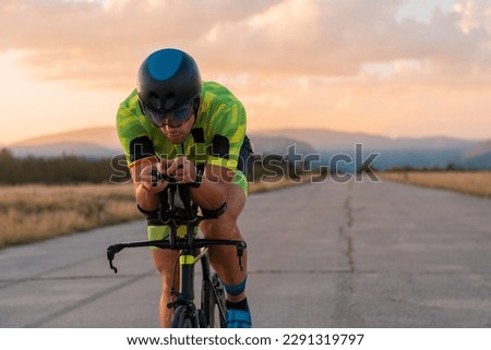  Triathlete riding his bicycle during sunset, preparing for a marathon. The warm colors of the sky provide a beautiful backdrop for his determined and focused effort. Royalty-Free Stock Photo #2291319797