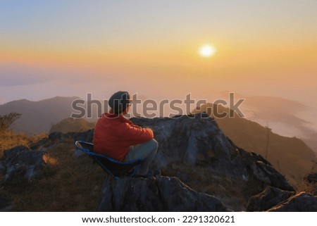 Portrait and landscape view of tourist wears red jacket and sit down on top of rock mountain with sunrise and fog background, Doi Pha Mon, Chiang Rai, Thailand
