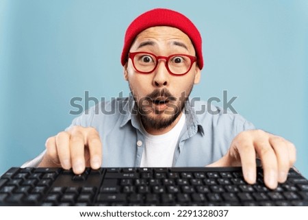 Amazed asian man wearing red hat, eyeglasses typing on keyboard, shopping online isolated on blue background. Emotional Korean hipster playing game. Portrait of emotional programmer working project