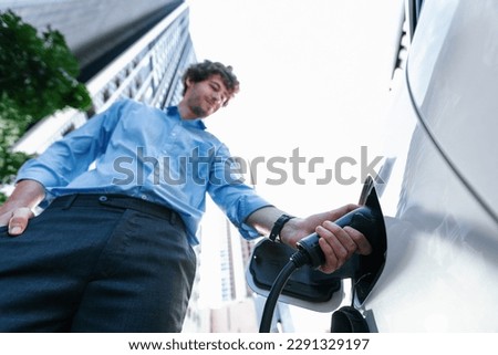Fisheye view focus on hand insert EV charger plug into electric car with blur background of progressive modern city lifestyle-businessman recharge his EV car with residential building condo background