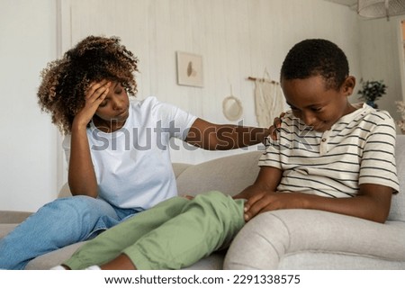 Exhausted African American mother grabs head from antics of kid boy who smiles maliciously. Naughty mischievous mixed race child son indulges while sit on couch unnerving ignore upbringing tired woman Royalty-Free Stock Photo #2291338575
