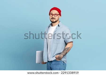 Smiling smart asian student holding laptop computer isolated on blue background, education concept. Portrait of Korean freelancer with stylish tattoo, wearing red hat, eyeglasses looking at camera