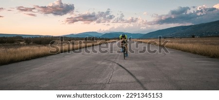  Triathlete riding his bicycle during sunset, preparing for a marathon. The warm colors of the sky provide a beautiful backdrop for his determined and focused effort. Royalty-Free Stock Photo #2291345153