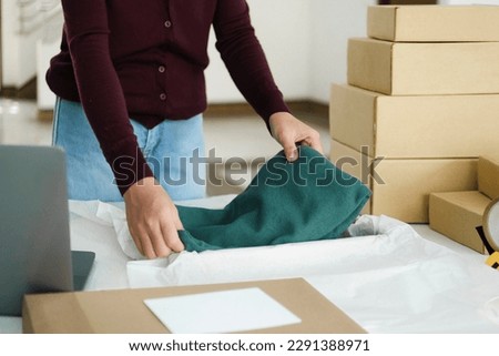 Close up of young female online business owner standing at table with cardboard boxes and laptop in workplace folding clothes, product into box for online order preparing for shipping and delivery.