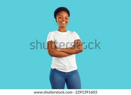 Studio shot of a happy, smiling woman in casual clothes. Portrait of a beautiful young African American girl in a white T shirt and jeans standing with her arms crossed isolated on a blue background Royalty-Free Stock Photo #2291391603