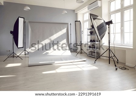 Photo session studio. Photographer's workplace in commercial atelier agency. Sunny workspace interior with grey backdrop, backstage and lamp, umbrella, flashlight illumination equipment kit. No people Royalty-Free Stock Photo #2291391619