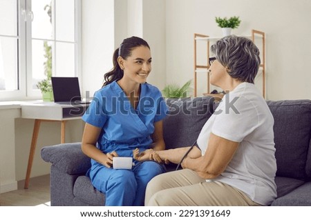 Smiling nurse measuring pressure of positive elderly woman. Caregiver visiting senior woman at home. Friendly nurse and patient talking during health check up. Caregiving, nursing of elderly people
