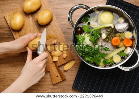 Woman peeling potato for cooking tasty bouillon at wooden table, top view Royalty-Free Stock Photo #2291396449