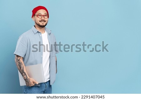 Portrait of smiling Korean freelancer with stylish tattoo, wearing red hat, eyeglasses looking at camera. Smart asian student holding laptop computer isolated on blue background, education concept