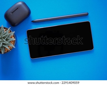 Top view, blank black screen of smartphone and wireless earphone in- charging case, pen on blue background. Template, mock-up empty screen of mobile phone for text and logo of product.