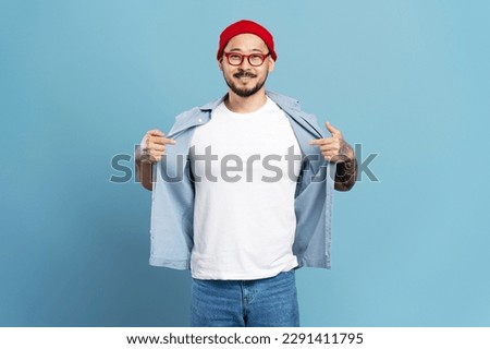 Portrait of smiling confident asian man, hipster wearing red hat, stylish eyeglasses, white t shirt pointing himself isolated on blue background. Mockup 