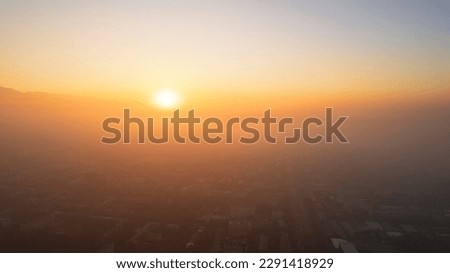 Epic gray smog is visible at sunset over the city. A bird's-eye view from a drone of houses, roads, cars and parks. White clouds and snowy mountains are illuminated by orange rays of the sun. Almaty Royalty-Free Stock Photo #2291418929