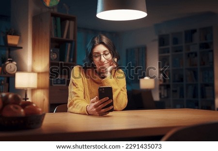 Young happy woman sitting in the living room at night and chatting with her smartphone, lifestyle and technology concept Royalty-Free Stock Photo #2291435073