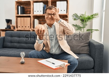 Senior psychiatrist man working at consultation office smiling happy doing ok sign with hand on eye looking through fingers 