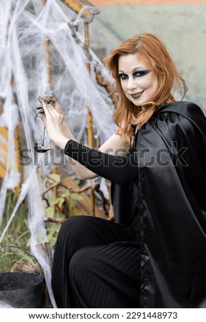 Attractive beautiful woman in a Halloween costume black cloak and scary make-up. Young girl in a look of a witch dressed up for a party making fairy potion holding black lantern. High quality photo