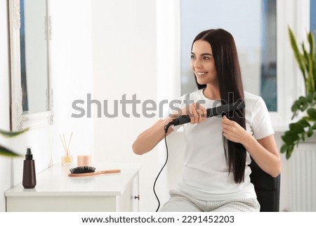 Beautiful happy woman using hair iron near mirror in room
